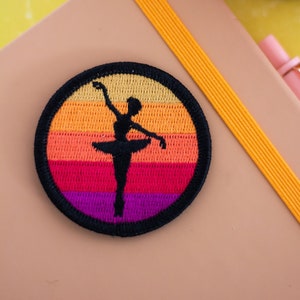 May include: A round embroidered patch featuring a black silhouette of a ballerina dancing against a multi-coloured striped background. The stripes are yellow, orange, red, pink and purple.