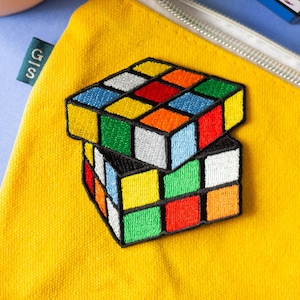 May include: Yellow canvas pouch with a silver zip and a brown leather pull tab. The pouch has an embroidered Rubik's Cube patch. A partially visible Rubik's Cube sits on a blue surface next to the pouch. A small green tag with the letters "G|S" is attached.