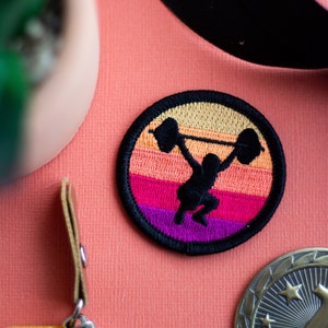 May include: A black and white embroidered patch featuring a silhouette of a person lifting a barbell against a sunset backdrop. The sunset is represented by four horizontal stripes in shades of orange, pink, and purple. The patch is circular and has a black border.