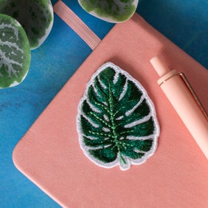 以下が含まれることがあります： ピンクのノートに緑色の刺繍の葉のパッチとピンクのペンが置かれている