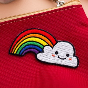Puede incluir: Una bolsa roja con cremallera con un parche bordado de un arcoíris y una nube. La nube tiene una cara sonriente.