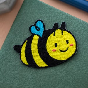 Puede incluir: Un parche bordado de abeja con un cuerpo rayado amarillo y negro, un ala de corazón azul y una cara sonriente con mejillas naranjas. El parche está sobre un fondo verde.