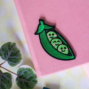 May include: A green pea pod with three smiling peas inside. The pod is embroidered with a light pink blush on each pea. The patch is on a pink background.