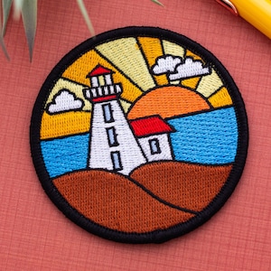 Op de afbeelding: Gestikte patch met een witte vuurtoren met een rood dak, tegen een kleurrijke zonsondergang boven een blauwe zee. De patch is rond en heeft een zwarte rand.