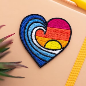 Puede incluir: Un parche bordado en forma de corazón con una ola azul y una puesta de sol en tonos naranja, rojo y rosa. El sol es amarillo y blanco.