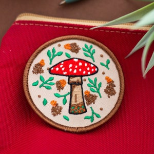 May include: A red and white mushroom with white spots is embroidered on a beige fabric patch. The patch is surrounded by green leaves and brown pine cones. The patch is attached to a red fabric pouch.