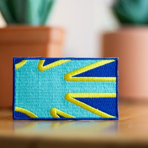 May include: A blue and yellow embroidered patch with a geometric design. The patch is square and has a soft, fuzzy texture.