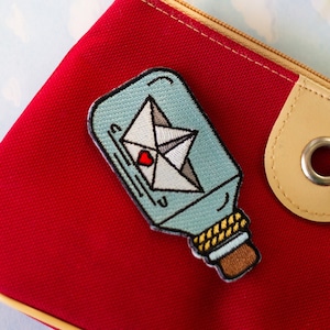Puede incluir: Una bolsa roja con cremallera que tiene un parche bordado azul y blanco de un mensaje en una botella. El mensaje es un papel doblado con un corazón y un pequeño velero dentro de la botella.