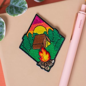 Peut inclure: Patch brodée thermocollante représentant une scène de camping avec une tente, un feu de camp et un coucher de soleil. Le patch est vert, marron, orange, jaune et rose.