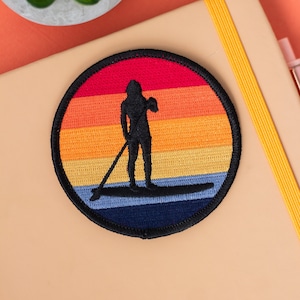 Pode incluir: Um remendo redondo bordado com uma silhueta de uma pessoa fazendo stand-up paddleboarding em frente a um pôr do sol colorido. O remendo tem um contorno preto e é costurado em uma superfície bege.
