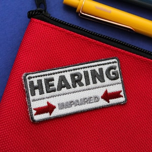 Puede incluir: Una bolsa roja con cremallera con una cremallera negra y un parche blanco y gris que dice "HEARING IMPAIRED" con una flecha roja apuntando hacia la derecha.