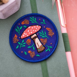 Op de afbeelding: Een blauwe en paarse geborduurde patch met een rode en witte paddenstoel omringd door groene bladeren en oranje bloemen.