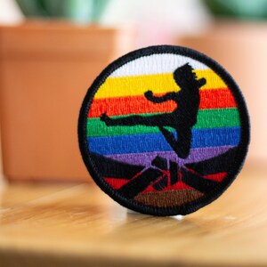 Puede incluir: Un parche redondo bordado con un fondo arcoíris y una silueta de una persona dando una patada en pose de karate. El parche tiene un borde negro y un fondo marrón.