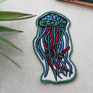 Puede incluir: Un parche bordado de medusa azul y rojo, con fondo blanco y contorno verde. El parche está sobre una superficie gris.