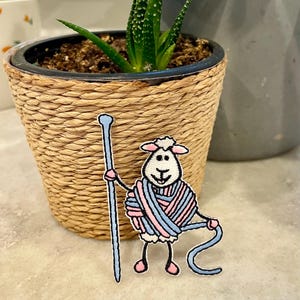 May include: A white sheep with pink cheeks is holding a blue knitting needle and is wrapped in pink and blue yarn. The sheep is standing on a light gray surface.