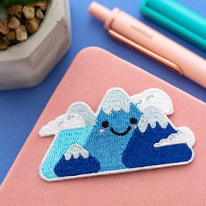 May include: A blue and white embroidered patch of a smiling mountain with clouds. The patch is on a pink notebook.