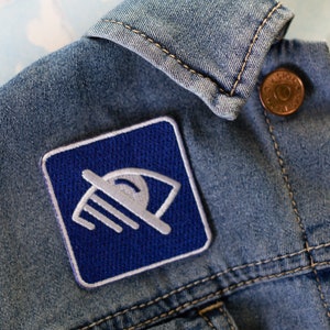 Puede incluir: Una chaqueta vaquera azul con un parche de un ojo blanco y azul con una barra diagonal cosido en el lado izquierdo delantero de la chaqueta.