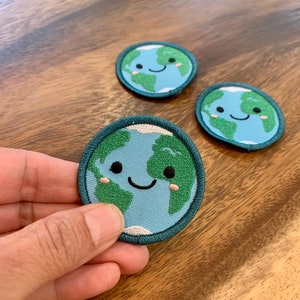 A hand holding an iron-on patch of the Earth with a cute smiling face and small pink cheeks with two patches in the background