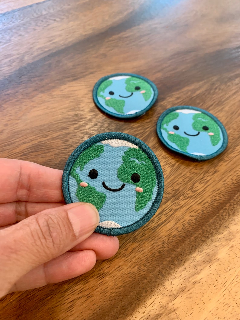 A hand holding an iron-on patch of the Earth with a cute smiling face and small pink cheeks with two patches in the background