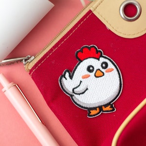 Può includere: Borsa rossa con cerniera con un designo di pollo ricamato bianco. Il pollo è rivolto a destra e ha una cresta e un becco rossi. La borsa ha una tiretta della cerniera beige e una patta in pelle beige con un anello di metallo.