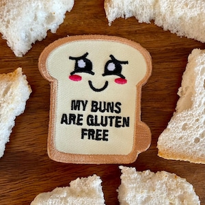 Könnte beinhalten: Ein gestickter Aufnäher in Form einer Brotscheibe mit einem lächelnden Gesicht und dem Text "MY BUNS ARE GLUTEN FREE". Der Aufnäher ist von Brotstücken auf einer Holzoberfläche umgeben.