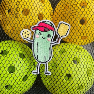 May include: A black mesh bag filled with yellow and green pickleballs. A patch of a cartoon pickle wearing a red visor and holding a paddle and a pickleball is in the center. The pickle is green with black details.