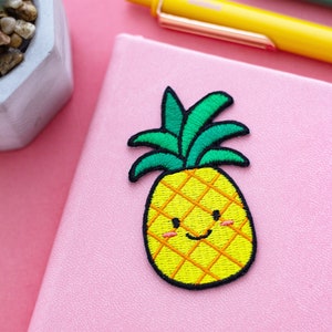 May include: A pink notebook with a yellow and green embroidered pineapple with a smiling face. The pineapple is a cute cartoon style.