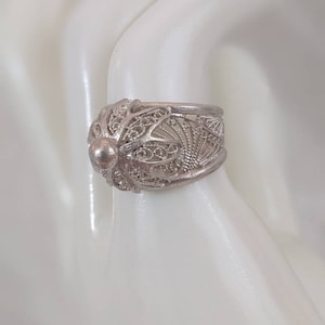 Può includere: Un anello in filigrana d'argento con un design ornato. L'anello presenta delicati motivi traforati e un dettaglio sferico centrale. L'anello è esposto su una superficie bianca, mettendo in mostra la sua intricata lavorazione artigianale e lo stile vintage.