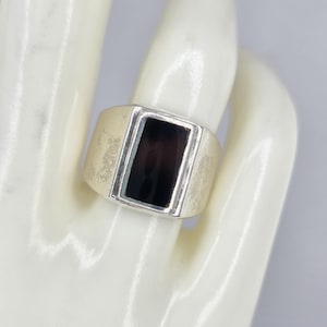 May include: A silver-toned ring with a rectangular black stone set in the center. The ring has a brushed metal finish and a thick band. The black stone is framed by a thin silver border.