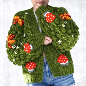 Può includere: Un cardigan verde lavorato a maglia con un motivo strutturato, con applicazioni ricamate di funghi rossi e bianchi, foglie arancioni e gialle e accenti di margherite bianche. Il cardigan ha maniche lunghe e una vestibilità ampia.