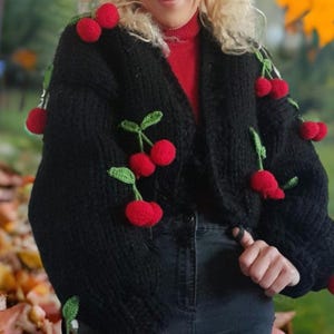 Puede incluir: Un cárdigan negro de punto grueso adornado con aplicaciones de cerezas rojas y hojas verdes. El suéter tiene cuello en V y mangas largas. Las cerezas están espaciadas uniformemente en la parte delantera y las mangas.