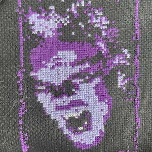 May include: Purple and white cross-stitch embroidery of a person's face on a black fabric background.
