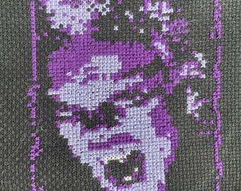 May include: Purple and white cross-stitch embroidery of a person's face on a black fabric background.