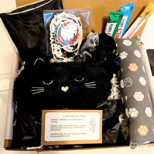May include: A gift box containing a black cat-shaped eye pillow, stickers, snacks, and a gray paw print patterned item. The eye pillow has embroidered details and a heart. A card with instructions is included.