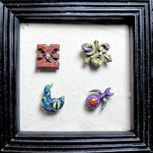 May include: Four small, hand-painted decorative emblems arranged in a square. One is a red block with a skull, one is a green and purple three-pointed star, one is a blue and green eye shape, and one is a purple sphere with a red center and silver blades.
