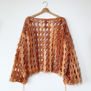 May include: A crocheted open-weave top in shades of orange and brown hangs on a wooden hanger against a white background. The top features a loose, flowing design with long sleeves and a boat neckline. The open weave creates a textured, airy look.