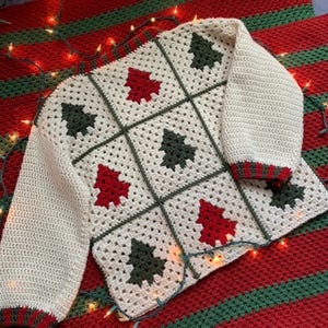 May include: A cream-colored, crocheted sweater featuring a grid pattern with red and green Christmas tree designs. The sweater has long sleeves with red and green striped cuffs, and is displayed on a red and green striped blanket.