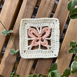 May include: A crocheted square with a pink butterfly design in the center. The square is made with white yarn and has a delicate, lacy look.