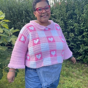 Pu&ograve; includere: Un maglione all'uncinetto rosa e bianco con un motivo a quadri vichy e cuori bianchi. Il maglione ha un colletto a volant e maniche 3/4.