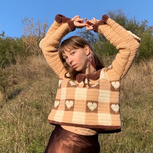 Pu&ograve; includere: Un maglione a uncinetto marrone e bianco con un motivo a forma di cuore e un colletto marrone. Il maglione ha un motivo a quadri ed &egrave; corto.