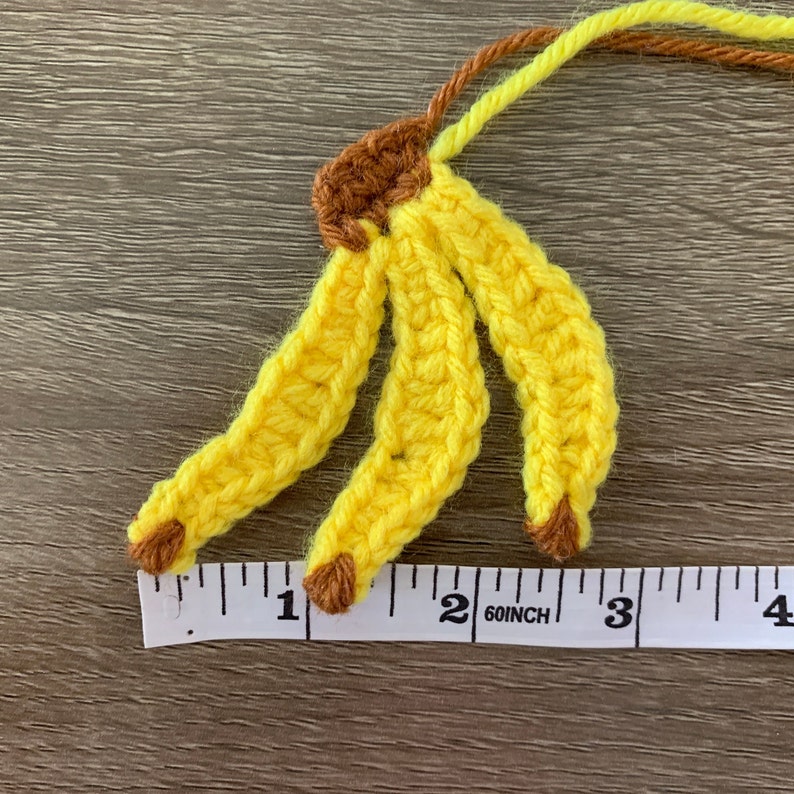Puede incluir: Un pl&aacute;tano de crochet hecho con hilo amarillo y marr&oacute;n. El pl&aacute;tano mide aproximadamente 5 cm de largo y se mide en una cinta m&eacute;trica blanca.