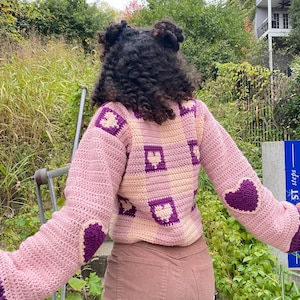 Pu&ograve; includere: Un maglione a uncinetto rosa e viola con un motivo a forma di cuore. Il maglione ha un design quadrato con cuori al centro di ogni quadrato. Il maglione &egrave; indossato da una persona con i capelli ricci.