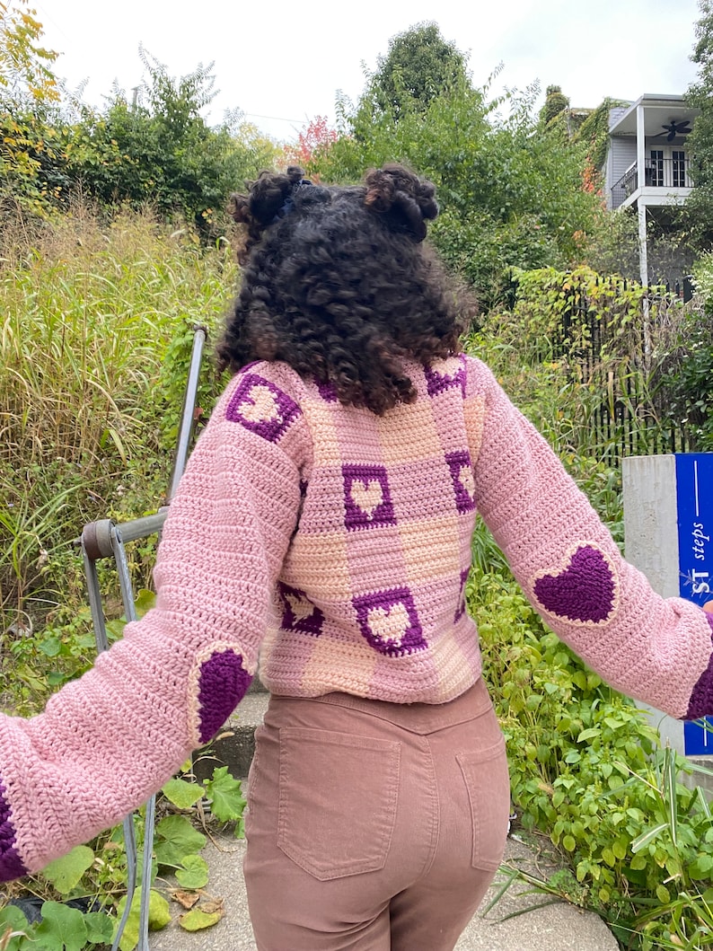 Pu&ograve; includere: Un maglione a uncinetto rosa e viola con un motivo a forma di cuore. Il maglione ha un design quadrato con cuori al centro di ogni quadrato. Il maglione &egrave; indossato da una persona con i capelli ricci.