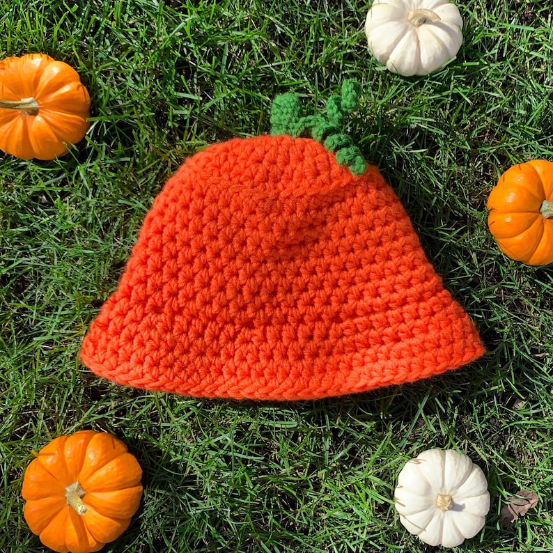 Pu&ograve; includere: Un cappello a forma di zucca di colore arancione lavorato a uncinetto con foglie verdi sulla cima. Il cappello &egrave; appoggiato su un letto di erba verde con diverse piccole zucche arancioni e bianche intorno.