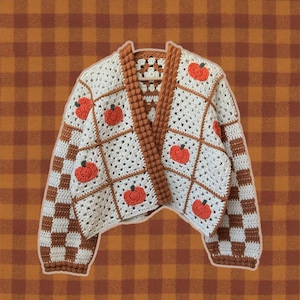 Può includere: Un cardigan all'uncinetto con un corpo a piastrelle granny color crema decorato con motivi a zucca arancioni. Le maniche e le rifiniture presentano un motivo a scacchi marrone e crema, e i bordi anteriori sono rifiniti con un bordo marrone testurizzato.