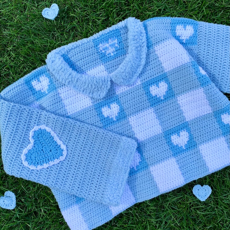 Pu&ograve; includere: Un maglione di uncinetto blu chiaro con un motivo a scacchi bianco e blu chiaro e dettagli a forma di cuore. Il maglione ha un colletto alla Peter Pan e maniche lunghe.