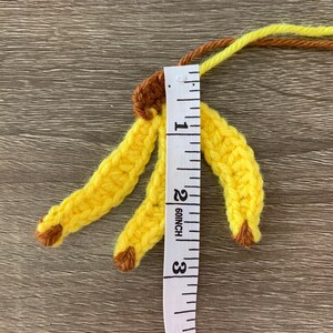 Puede incluir: Dos pl&aacute;tanos amarillos de crochet con puntas marrones, medidos con una cinta m&eacute;trica blanca, mostrando que miden aproximadamente 5 cm de largo.