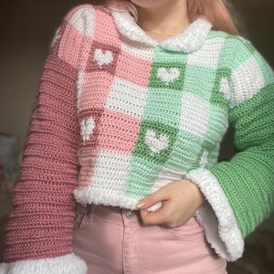 Pu&ograve; includere: Un maglione all'uncinetto rosa e verde con cuori bianchi e un colletto all'uncinetto bianco.