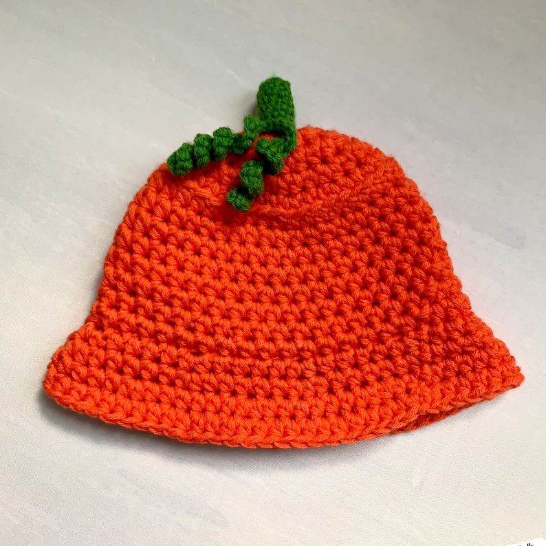 Pu&ograve; includere: Un cappello a forma di zucca lavorato a uncinetto di colore arancione con un gambo verde in cima.
