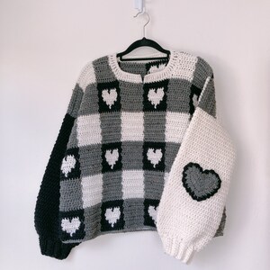 Pu&ograve; includere: Un maglione a uncinetto grigio e bianco con accenti a forma di cuore neri. Il maglione ha un motivo a scacchi e una vestibilit&agrave; ampia.